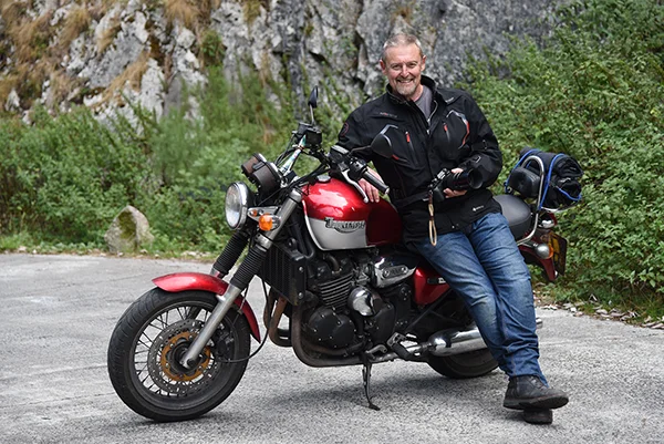 Photographer Mike Browne sitting on his Triumph motorcycle holding a camera